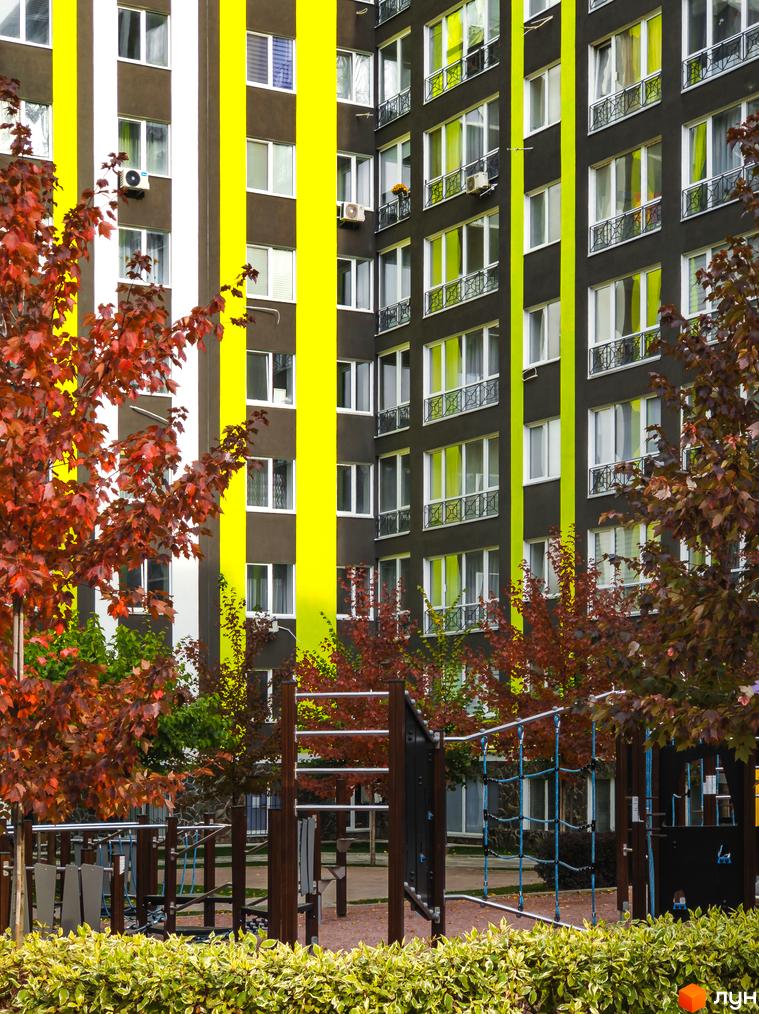 Висотна будівля ЖК Фортуна-2 з яскравими жовтими смугами на фасаді. На передньому плані дитячий майданчик з деревами з червоним листям.