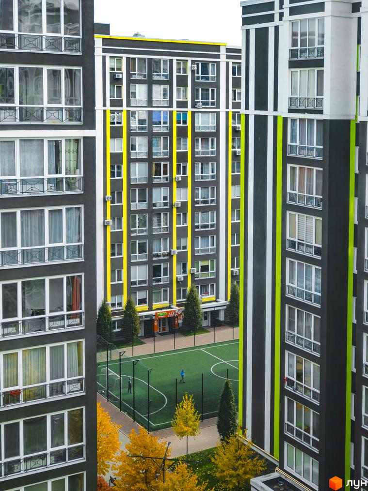 Високоповерхові будинки ЖК Фортуна-2 з фасадами чорного кольору з білими та жовтими смугами. На території є футбольне поле та дерева з жовтим листям.