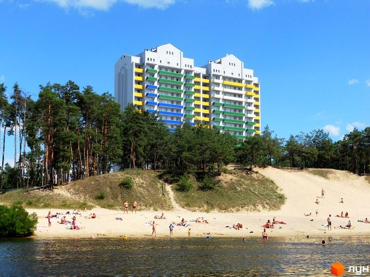 Житловий комплекс Райдужний з кольоровими балконами на фоні піщаного пляжу та лісу. Прибудинкова територія з відпочивальниками на пляжі.