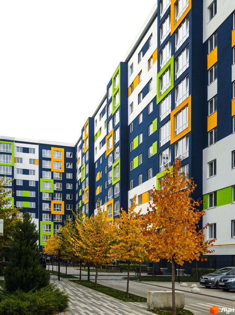 Житловий комплекс Модуль – яскрава новобудова з різнокольоровими фасадами та вікнами. Навколо будинку зелена територія з деревами та доглянутою доріжкою.