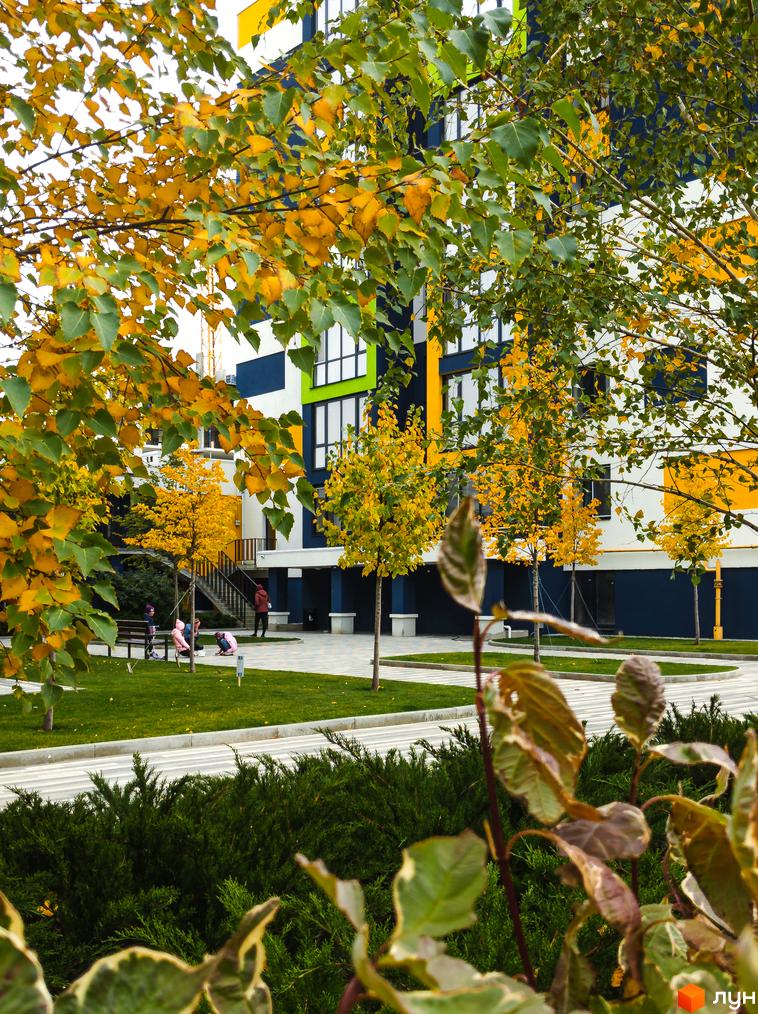 Житловий комплекс Модуль з яскравим фасадом у білих, синіх, жовтих та зелених кольорах. Прибудинкова територія з осінніми деревами, зеленим газоном та доріжками, де гуляють люди.