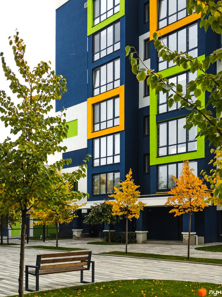 Новобудова Модуль: багатоповерховий будинок з синім, зеленим та жовтим фасадом. Облаштована територія з осінніми деревами, лавкою та мощеними доріжками.