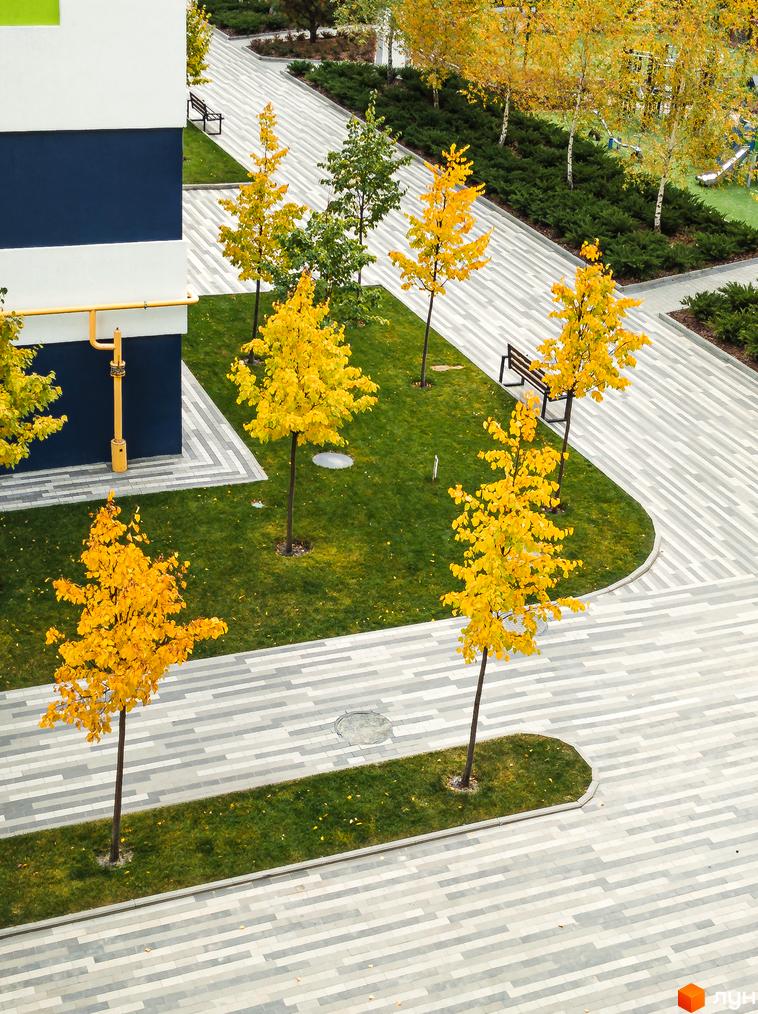 Житловий комплекс Модуль. Фасад будинку з синіми та білими елементами, поруч зелена зона з деревами та доріжками для прогулянок.