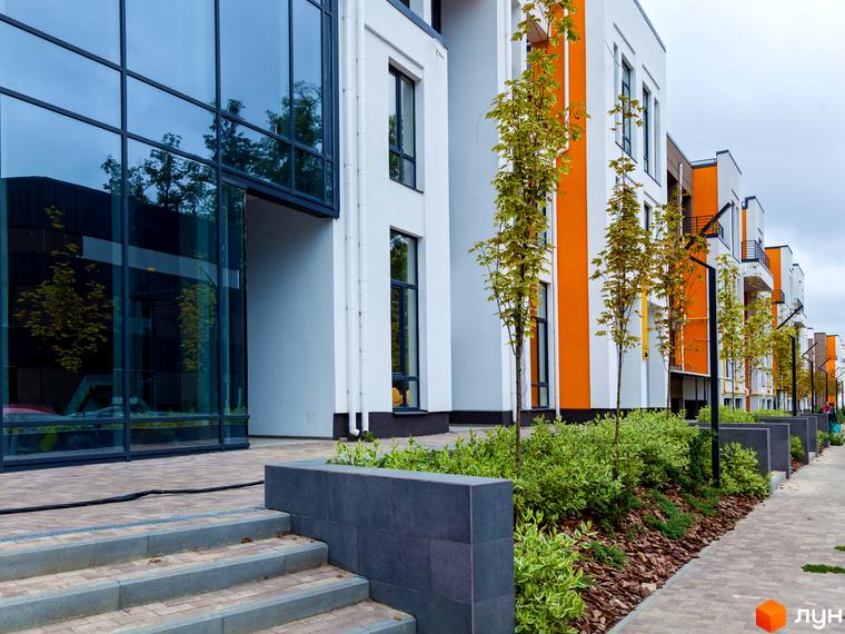 Новобудова Найкращий квартал: білі будинки з помаранчевими вертикальними вставками та скляним фасадом. Територія облаштована бруківкою, зеленими насадженнями та молодими деревами.