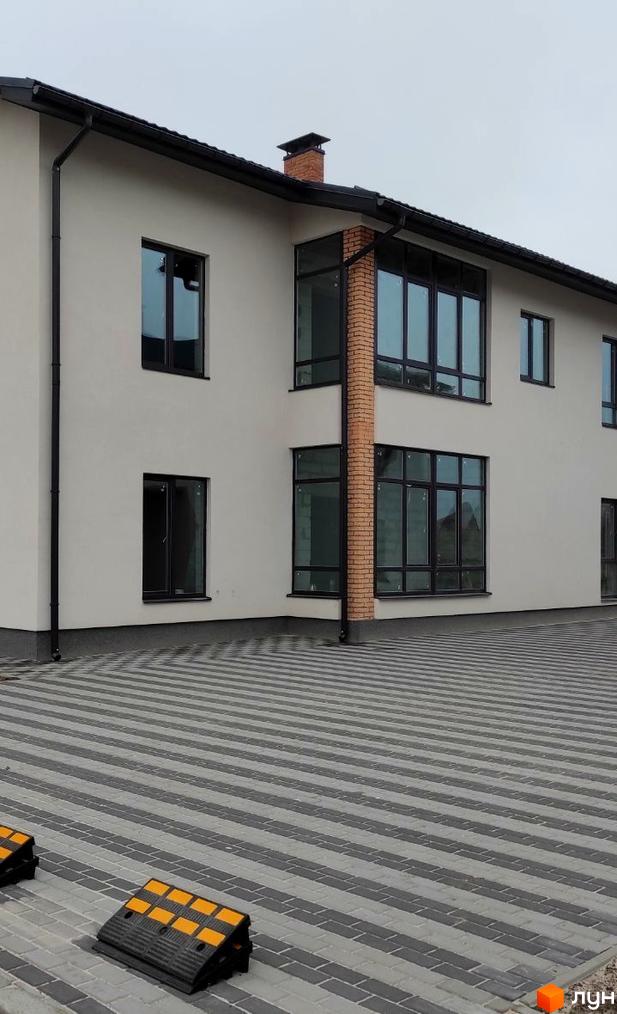 Білий двоповерховий будинок з великими вікнами та цегляним димарем. Територія перед будинком викладена сірою плиткою з паркувальними місцями.
