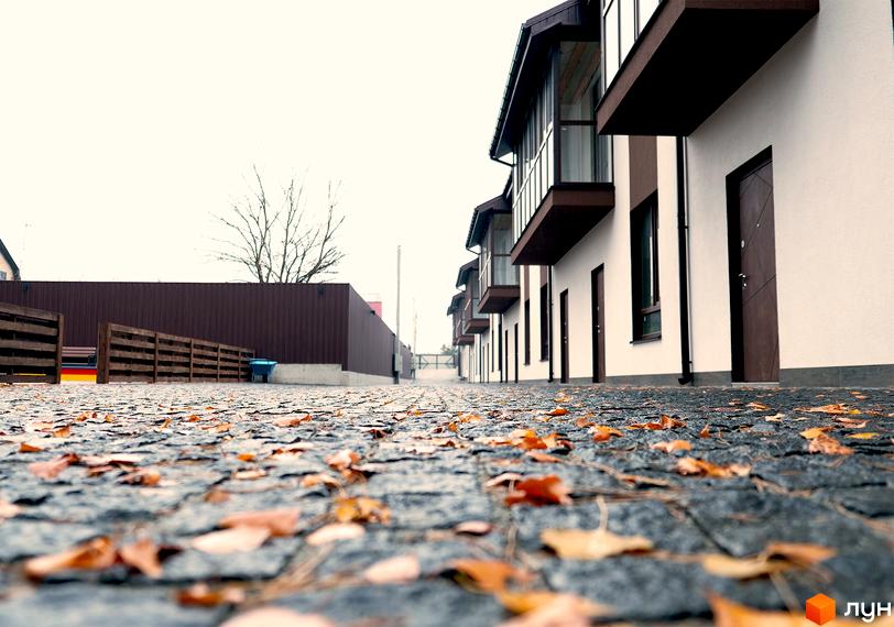 Ряд таунхаусів Таунхауси Green Heaven з білими фасадами та коричневими балконами. Територія вимощена бруківкою з опалим осіннім листям.