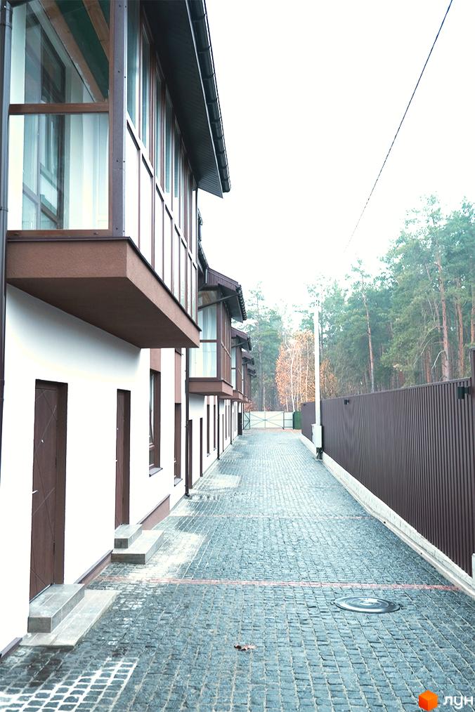 Зовнішній вигляд Таунхаусного комплексу Green Heaven: білі будинки з коричневими елементами та панорамними вікнами. Дорога з бруківки веде вздовж будинків до лісу.