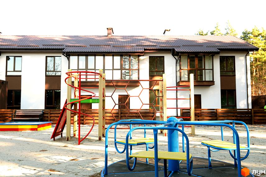 Фасад Таунхаусів Green Heaven: двоповерхові будинки з біло-коричневими стінами та коричневим дахом. На передньому плані дитячий майданчик з гіркою та каруселлю.