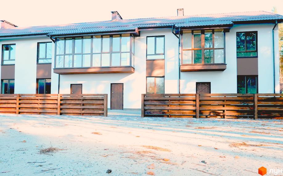 Фасад Котеджного містечка Green Heaven з білими стінами, коричневими вікнами та заскленими балконами. Перед будинком розташований дерев'яний паркан та ґрунтова ділянка.