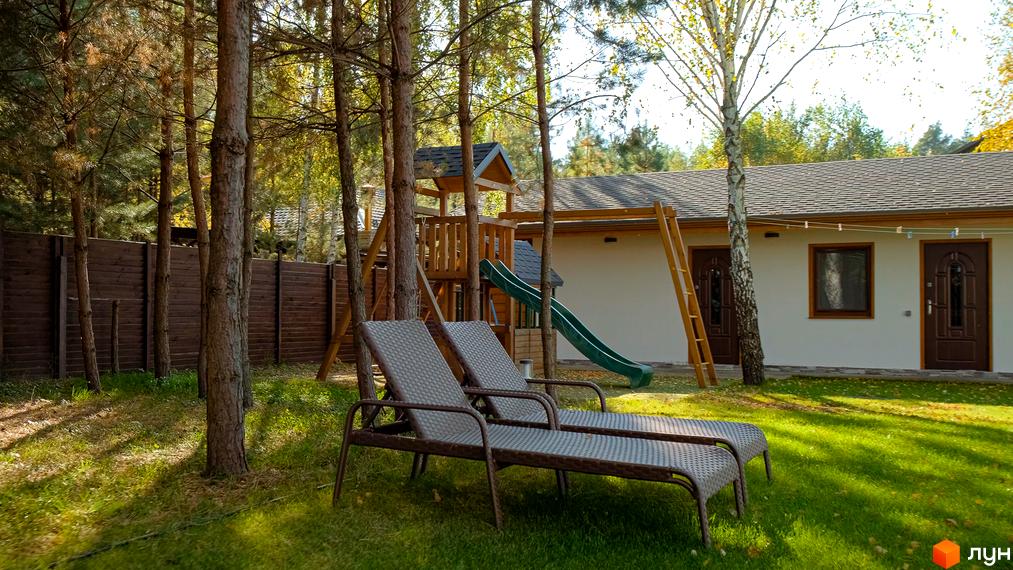 Котеджне містечко Калина-2: білий будинок з коричневими дверима та вікнами, поруч дитячий майданчик та шезлонги на зеленій галявині.