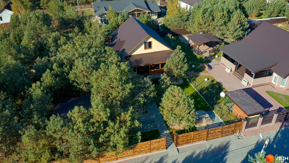 Котеджне містечко КМ Калина-2: двоповерховий будинок з коричневим дахом, оточений зеленими деревами, з доглянутою прибудинковою територією та дерев'яним парканом.