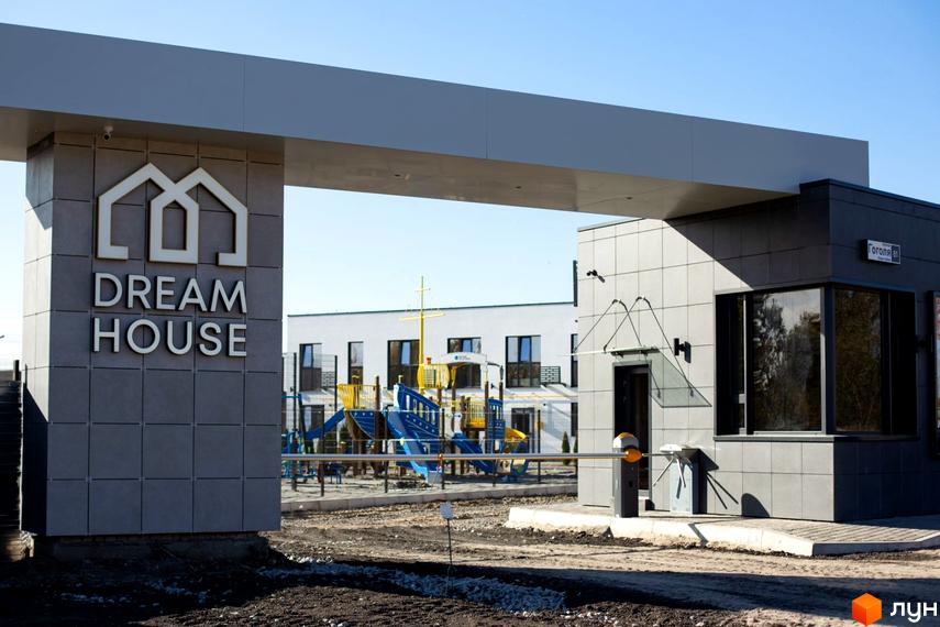 В'їзд до Таунхауси DREAM HOUSE з сірою пропускною будкою та шлагбаумом. На фоні видно білі будинки та дитячий майданчик.