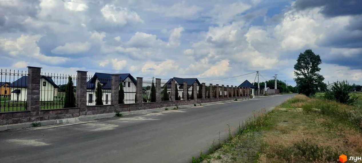 Котеджне містечко КМ Лелека: фасади будинків з темними дахами та білими стінами, огороджені парканом з кам'яними стовпами. Навколо - дорога та зелена територія.