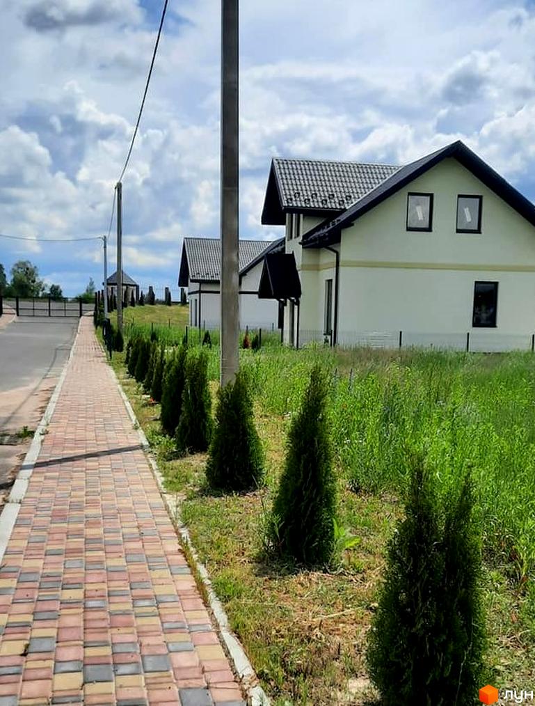 Будинок з двосхилим дахом, світлими стінами та темними вікнами. Перед будинком доглянута територія з тротуаром та зеленими насадженнями.