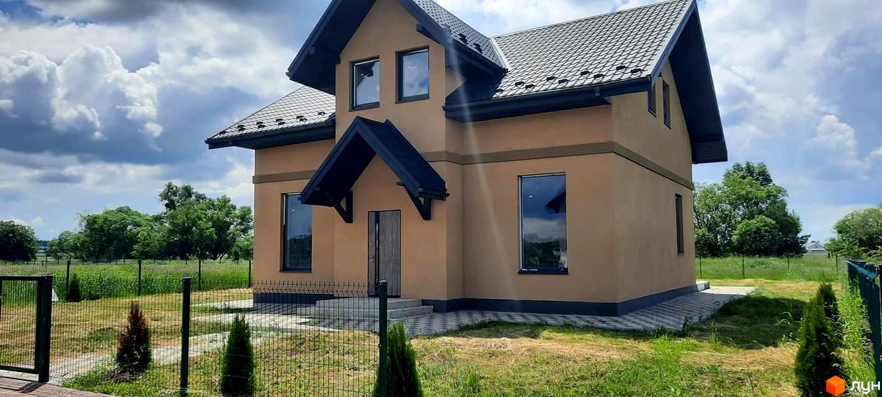 Двоповерховий будинок з темною покрівлею та вікнами, вхідним козирком. Перед будинком газон, огорожа та декоративні рослини.