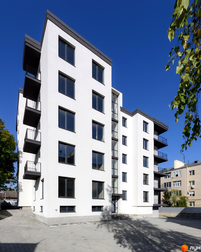 Фасад житлового комплексу на вул. Козачука, 27а, з балконами та великими вікнами на тлі синього неба. Прибудинкова територія вимощена плиткою.