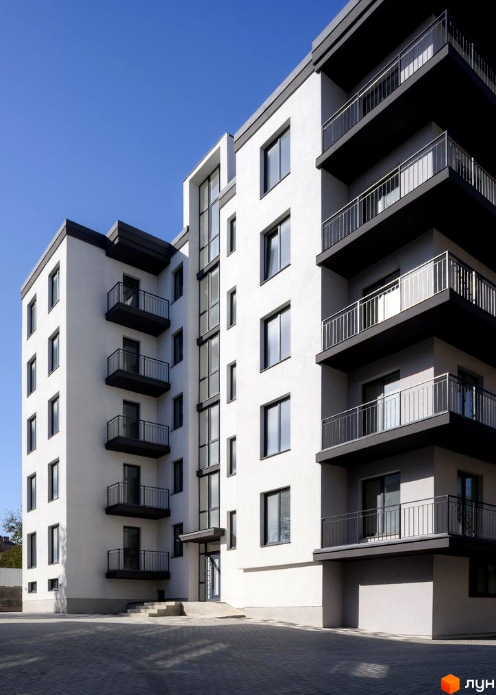 Зовнішній вигляд новобудови на вул. Козачука, 27а, з балконами та вікнами, на тлі блакитного неба. Прибудинкова територія з бруківкою.