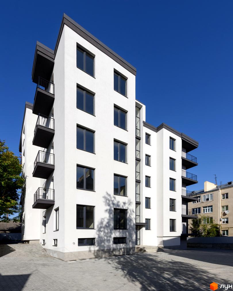 Новобудова на вул. Козачука, 27а. Фасад білої будівлі з темними балконами та вікнами, на тлі блакитного неба. Прибудинкова територія вимощена плиткою.