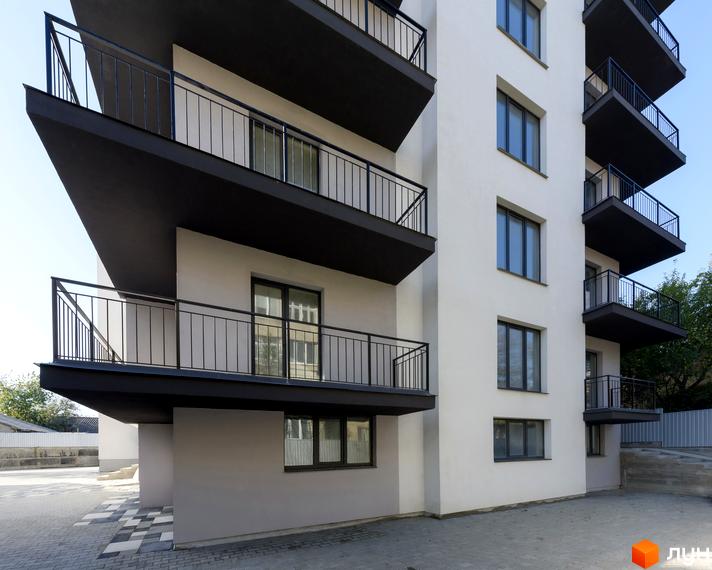 Фасад новобудови на вул. Козачука, 27а з балконами та вікнами на тлі блакитного неба. Прибудинкова територія з бруківкою та зеленими насадженнями.