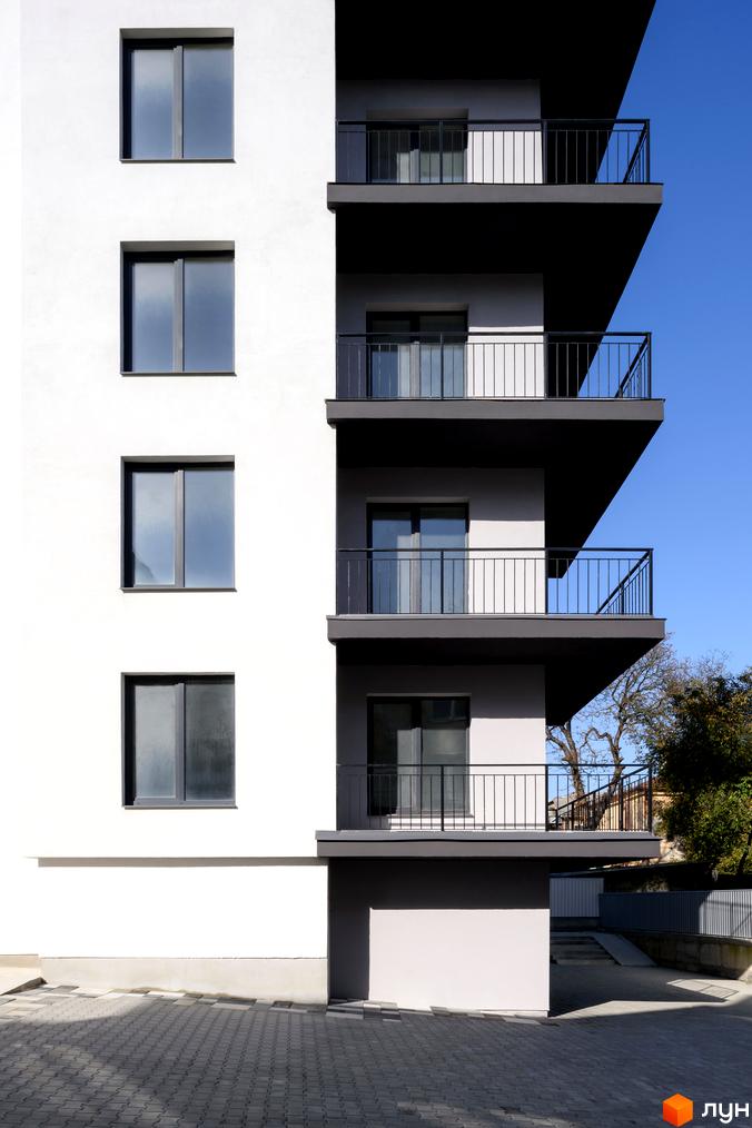 Зовнішній вигляд новобудови на вул. Козачука, 27а, з балконами та вікнами на тлі блакитного неба. Прибудинкова територія з бруківкою та деревами.