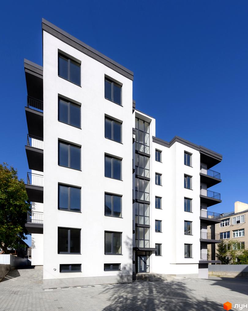 Зовнішній вигляд новобудови на вул. Козачука, 27а, з білими стінами, темними вікнами та балконами. Прибудинкова територія з бруківкою та деревами.