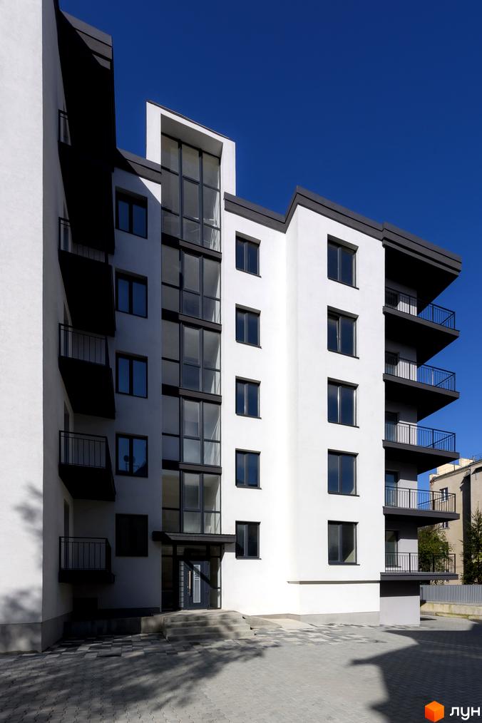 Зовнішній вигляд новобудови на вул. Козачука, 27а з балконами та великими вікнами на тлі блакитного неба. Прибудинкова територія з бруківкою та тінями від дерев.