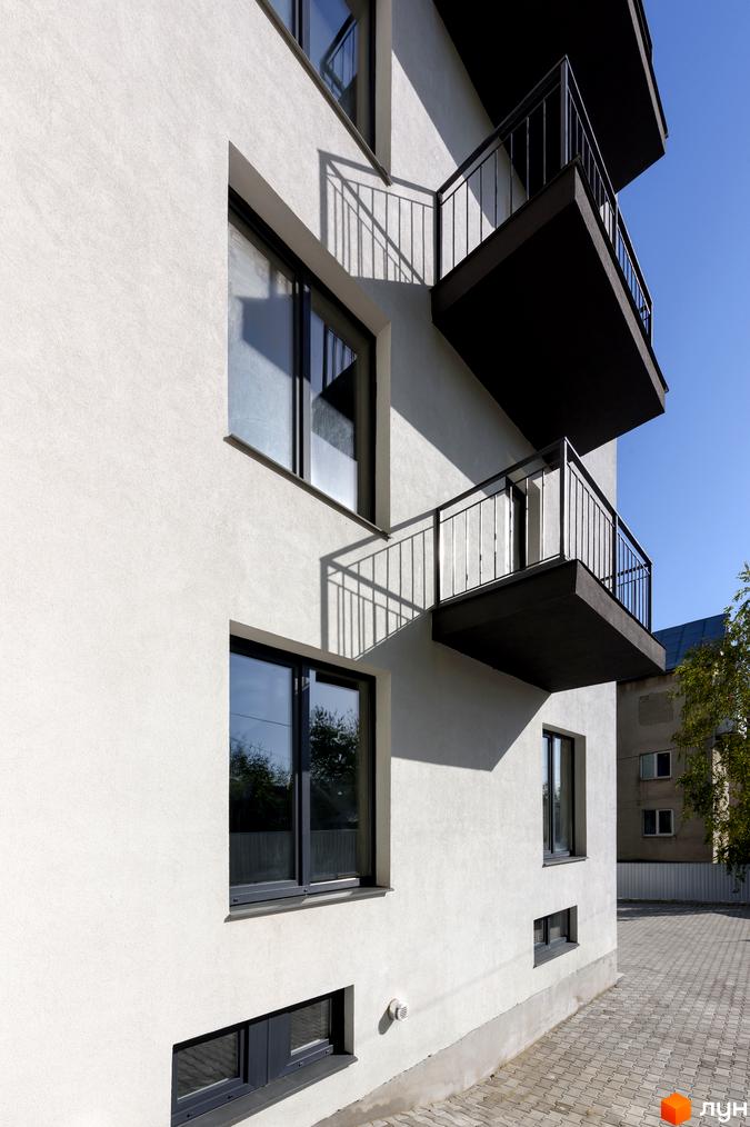 Фасад житлового комплексу вул. Козачука, 27а, з балконами та вікнами, на тлі блакитного неба. Прибудинкова територія з бруківкою та деревами.