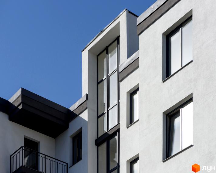 Фасад новобудови вул. Козачука, 27а, з великими вікнами та балконами на тлі блакитного неба. Зовнішній вигляд будинку з білою штукатуркою та темними елементами.