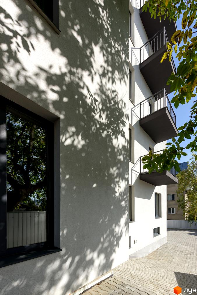 Зовнішній вигляд новобудови на вул. Козачука, 27а з балконами та тінями від дерев на стінах. Прибудинкова територія вимощена плиткою.