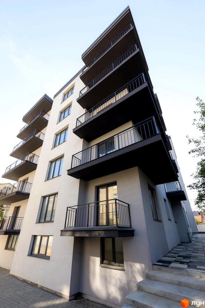 Зовнішній вигляд багатоквартирного будинку на вул. Козачука, 27а, з балконами та вікнами, на тлі блакитного неба. Територія перед будинком вимощена плиткою та має сходи.