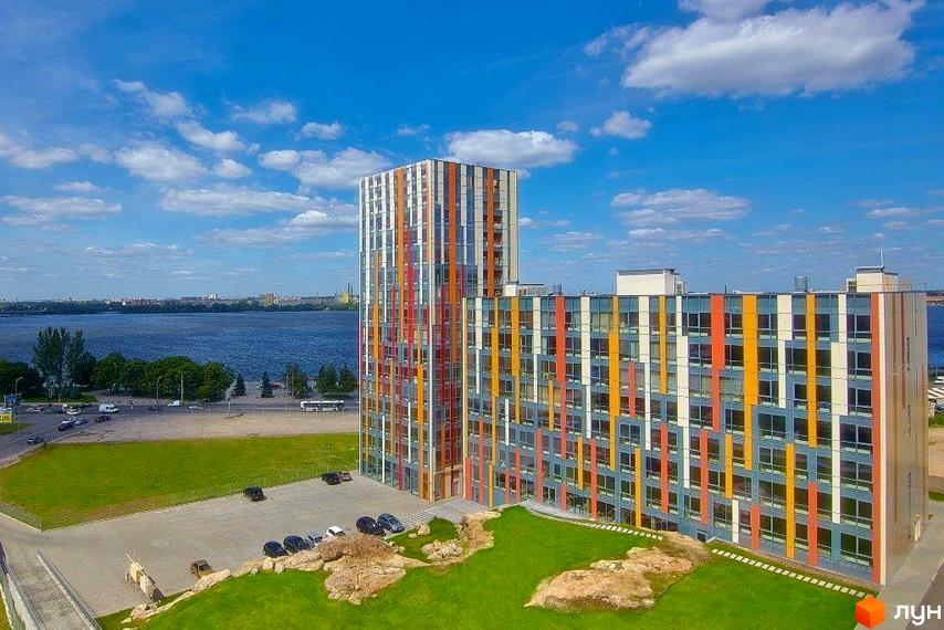 Житловий комплекс Фестивальний з кольоровими фасадами на березі річки. Навколо будівель зелені газони та парковка.