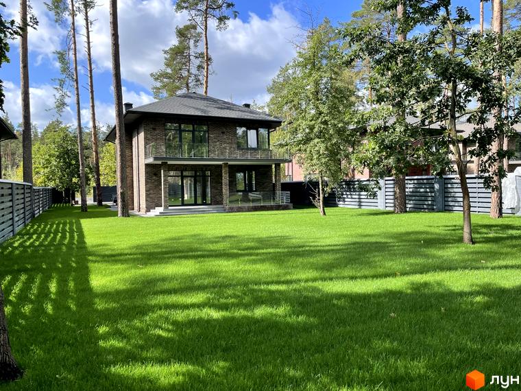 Котеджне містечко КМ Лісові Плюти - двоповерховий цегляний будинок з великими вікнами та доглянутою зеленою галявиною перед ним.