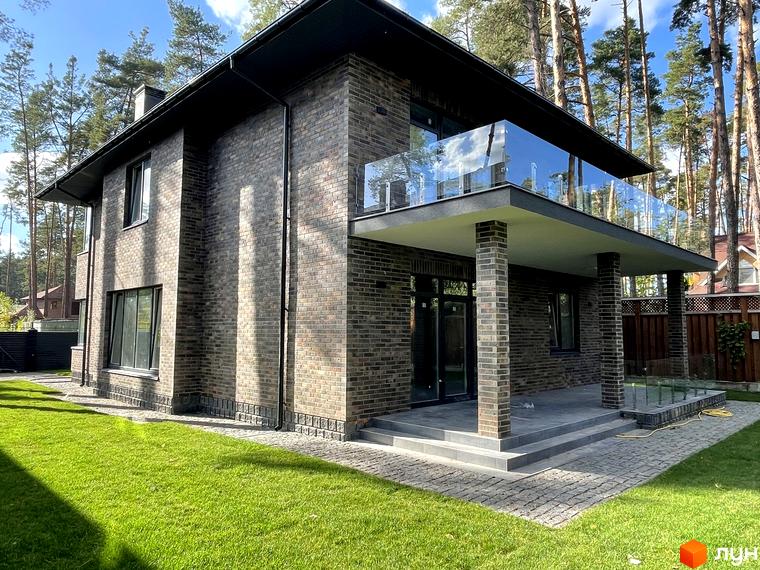 Котеджне містечко КМ Лісові Плюти. Будинок з цегляним фасадом, балконом зі скляним огородженням та доглянутою зеленою галявиною.