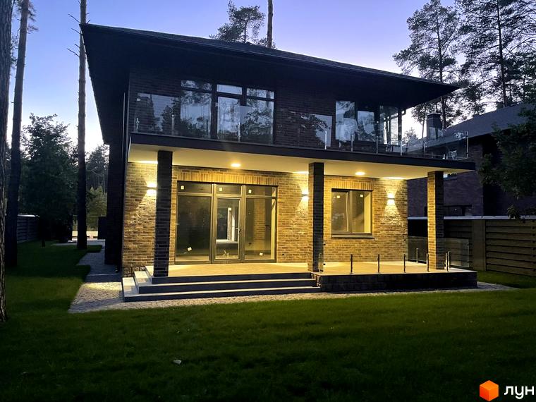 Котеджне містечко КМ Лісові Плюти. Зовнішній вигляд двоповерхового будинку з цегляним фасадом, великими вікнами та терасою. Прибудинкова територія з газоном та доріжками.