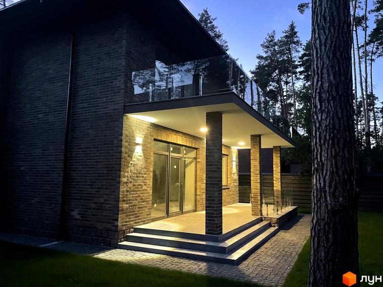 Котеджне містечко КМ Лісові Плюти: двоповерховий будинок з цегляним фасадом та скляним балконом, освітлена тераса та доріжка з бруківки.