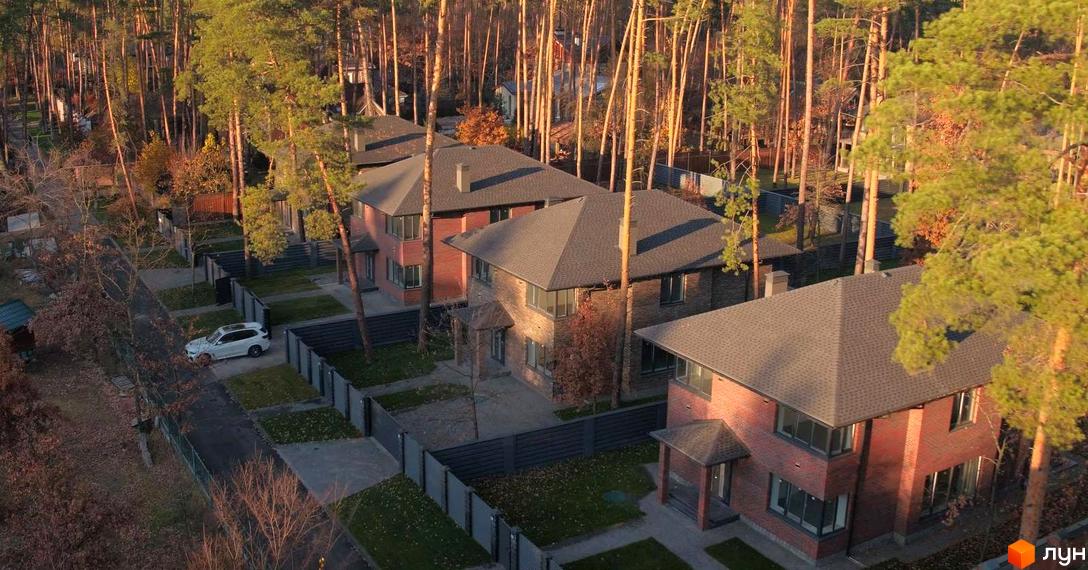 Котеджне містечко КМ Лісові Плюти: цегляні будинки з темними дахами, огороджена територія та зелені насадження навколо. На території комплексу розташовані приватні будинки та паркомісця.