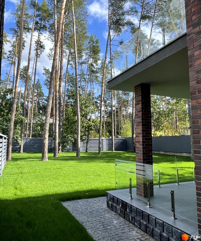 Котеджне містечко КМ Lisovi Plyuty з цегляним фасадом та скляним балконом на тлі соснового лісу. Доглянута прибудинкова територія з газоном та бруківкою.