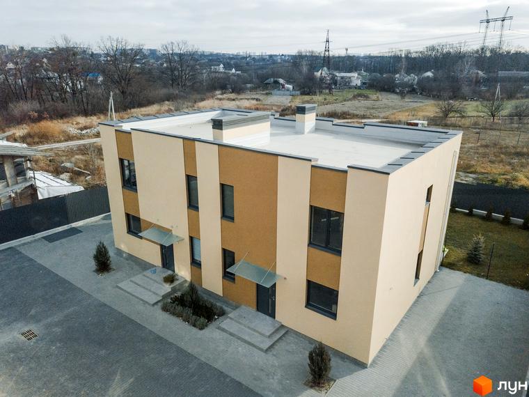 Зовнішній вигляд двоповерхового будинку котеджного містечка КМ My Land з сірим дахом та коричнево-бежевими фасадами, прибудинкова територія вимощена плиткою.