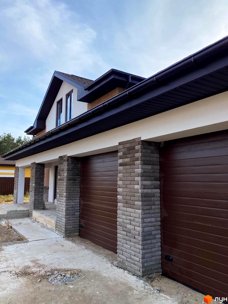 Котеджне містечко КМ Барвіта: фасади будинку з коричневими гаражними воротами та цегляними колонами. Прибудинкова територія з бетонною доріжкою та будівельним сміттям.