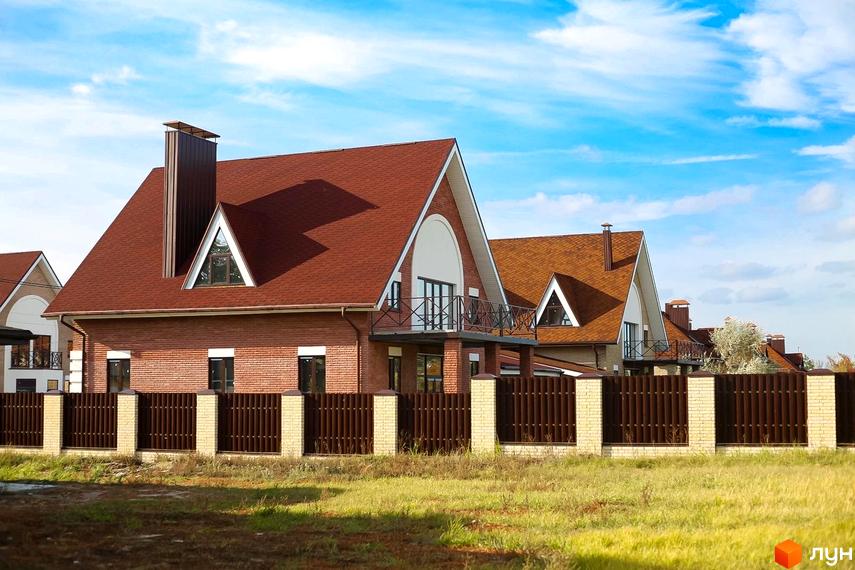 Котеджне містечко КМ Барвіта: цегляні будинки з червоними дахами та кованими балконами, огороджені коричневим парканом. Навколо зелена трава під блакитним небом.