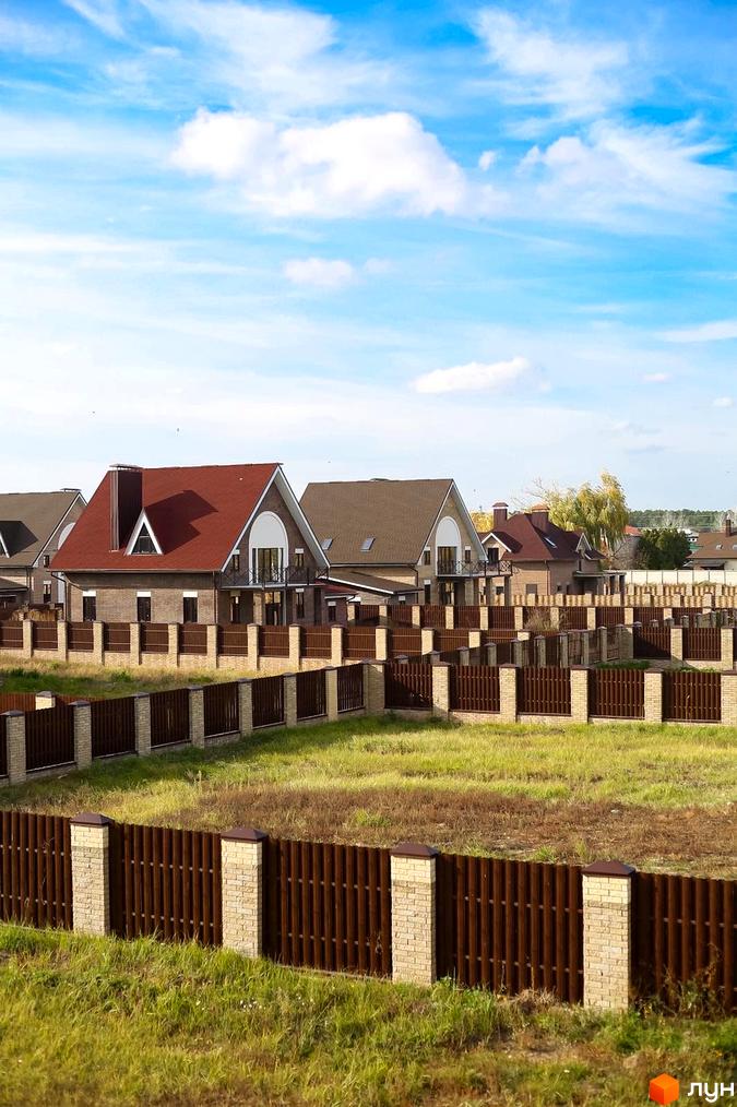Котеджне містечко КМ Барвіта: цегляні будинки з червоними дахами, огороджені парканом з цегляними стовпами. Навколо зелена трава та блакитне небо з хмарами.