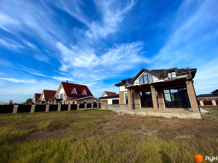 Котеджне містечко КМ Барвіта: двоповерхові будинки з червоної цегли з великими вікнами та балконами на тлі блакитного неба з хмарами. Навколо будівель - зелений газон та паркан.