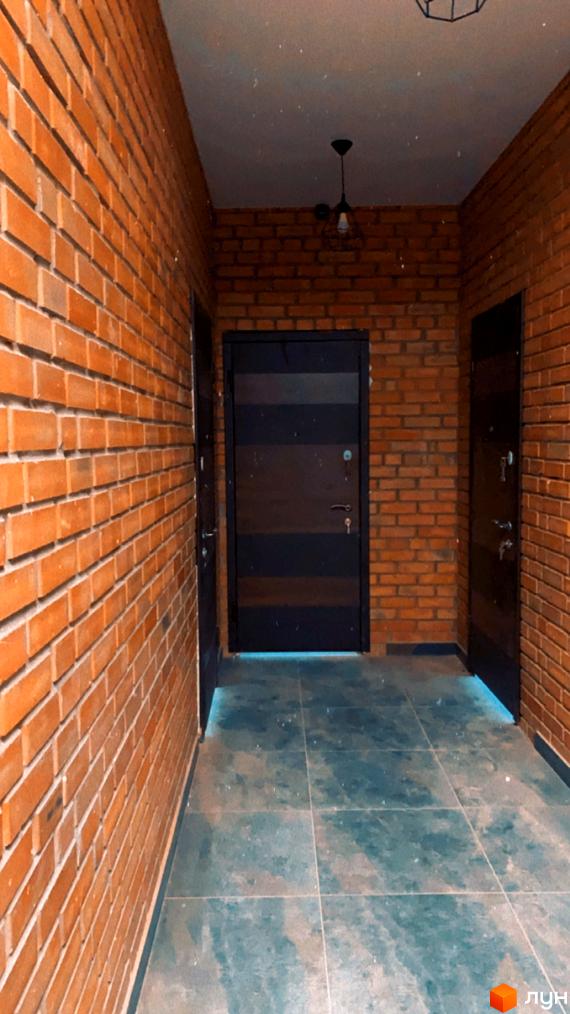 Інтер'єр коридору в Житловому комплексі Loft з цегляними стінами та темними дверима. На підлозі плитка, а вгорі видно світильник.