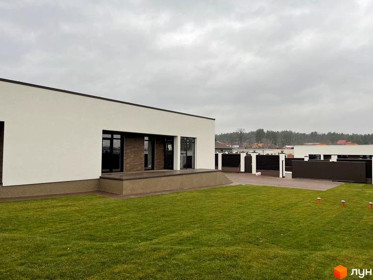 Котеджне містечко КМ White Stone: одноповерховий будинок з великими вікнами та терасою на тлі зеленого газону та огородженої території.