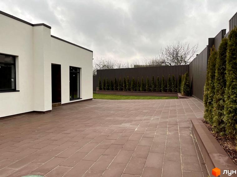 Зовнішній вигляд будинку КМ White Stone з білою стіною та коричневою плиткою на подвір'ї. Навколо будинку розташований паркан та зелені насадження.