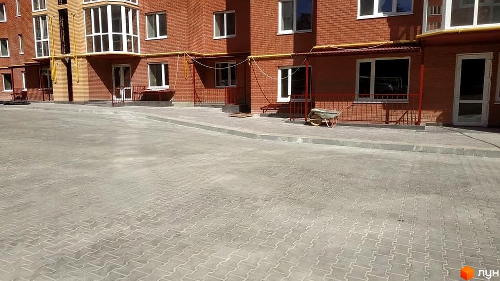 Цегляний житловий будинок з білими вікнами та балконами, жовтими трубами на фасаді. Прибудинкова територія вимощена сірою бруківкою.