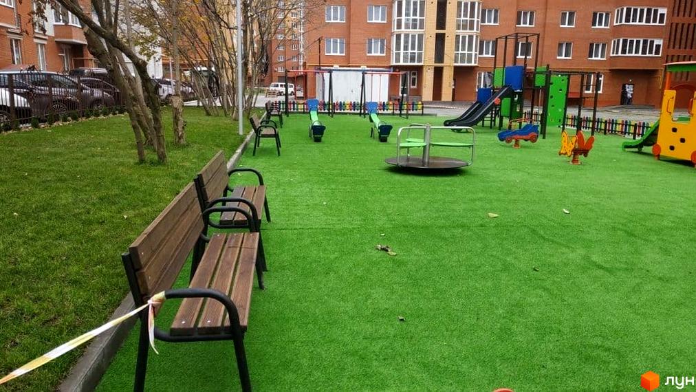 Зображення дитячого майданчика біля цегляного будинку. На території є гойдалки, карусель та лавки для відпочинку.