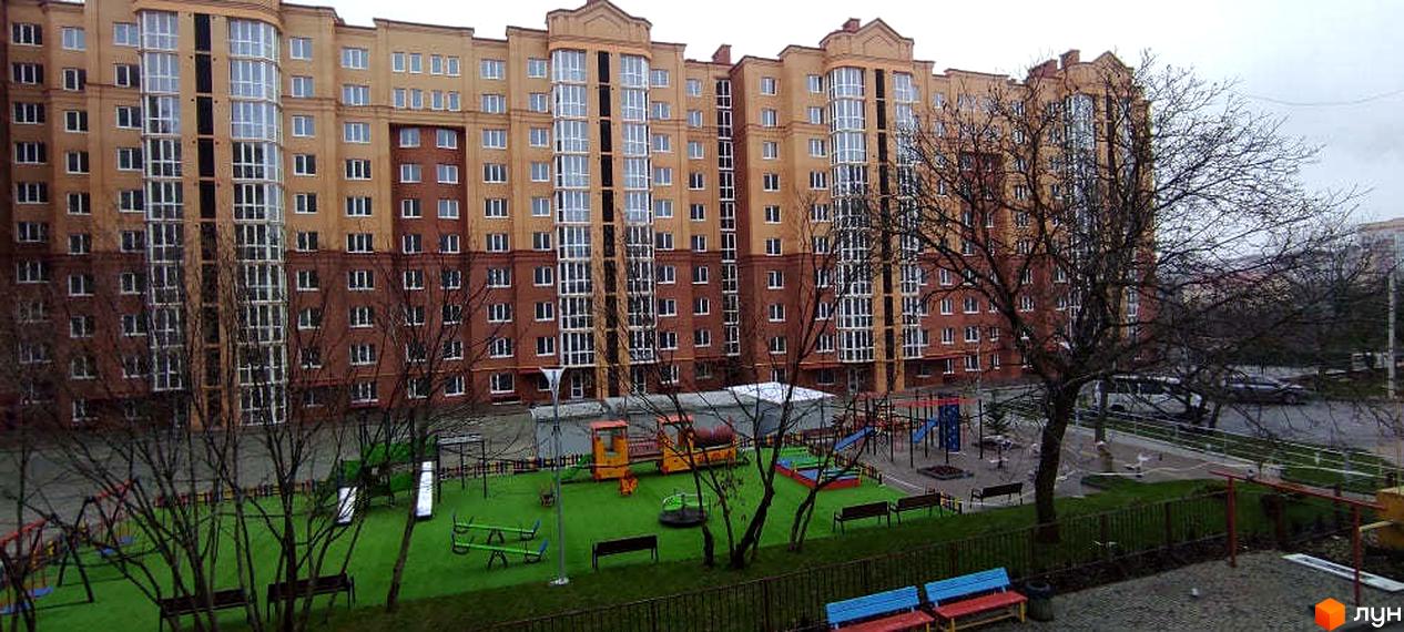 Житловий будинок з комбінованим фасадом та великими вікнами. На прибудинковій території облаштовано дитячий майданчик із зеленим покриттям.