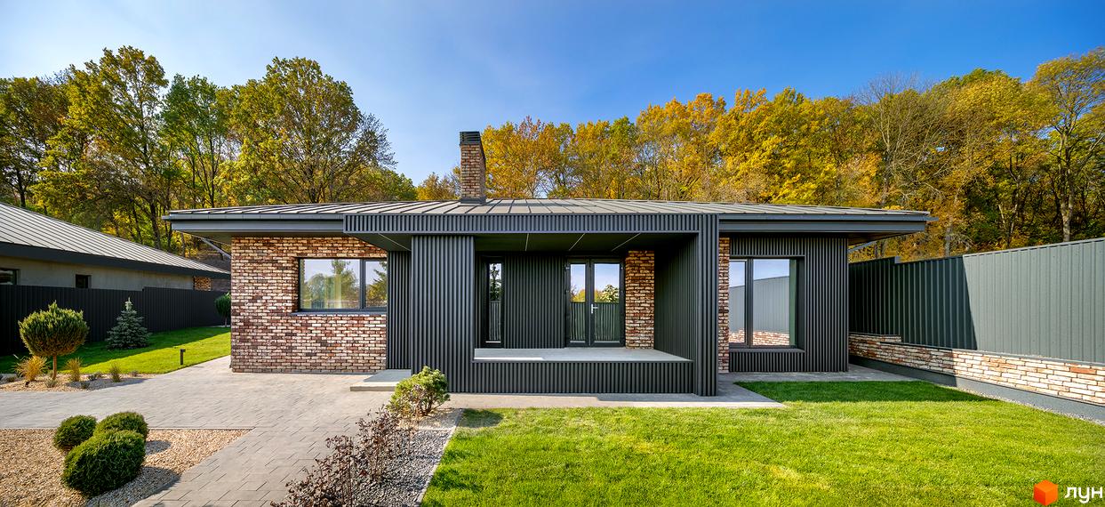 Одноповерховий будинок з цегляними стінами та темною покрівлею. На передньому плані газон та доріжка з бруківки, видно дерева на задньому плані.