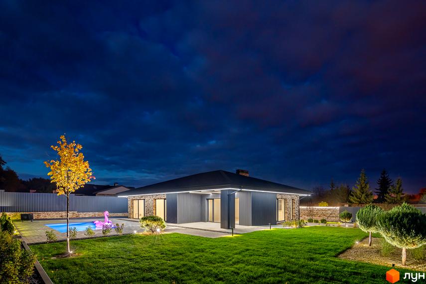 Освітлений будинок з темним фасадом та терасою. На території є газон, басейн з надувним фламінго та дерева.